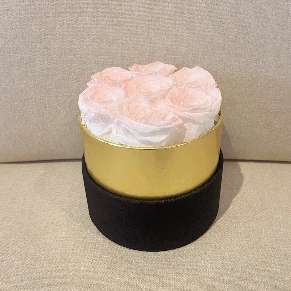 A round gift box containing pale pink roses wrapped in white tissue paper, with a shiny gold middle band and a matte black bottom band, set against a neutral beige textured background.