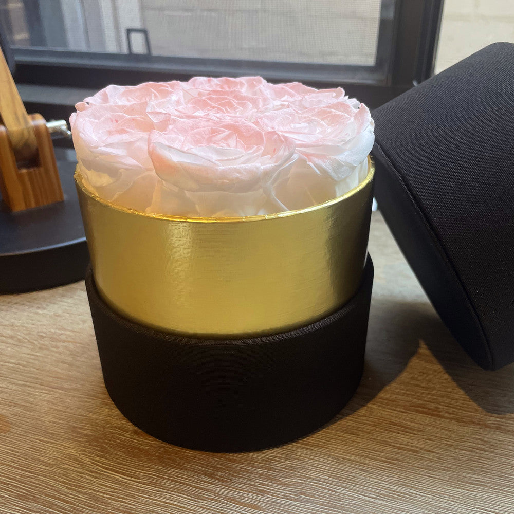 A round flower box with a black fabric base, a metallic gold middle band, and a lid leaning against it. The top is filled with pale pink preserved roses, photographed on a wooden tabletop near a window.