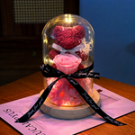 A decorative glass dome keepsake display on a wooden base, featuring a pink rose, red heart-shaped flower, pink petals, and warm string lights inside, tied with a black ribbon printed with 'Just for you'.