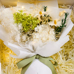 Close-up of Eternal Glow Preserved Hydrangea Gift Box interior showing a lush green and white preserved hydrangea bouquet with dried daisies, fluffy white pampas, and rosemary sprigs, wrapped in white floral paper with a sage green ribbon, nestled in cream shredded paper with warm fairy lights