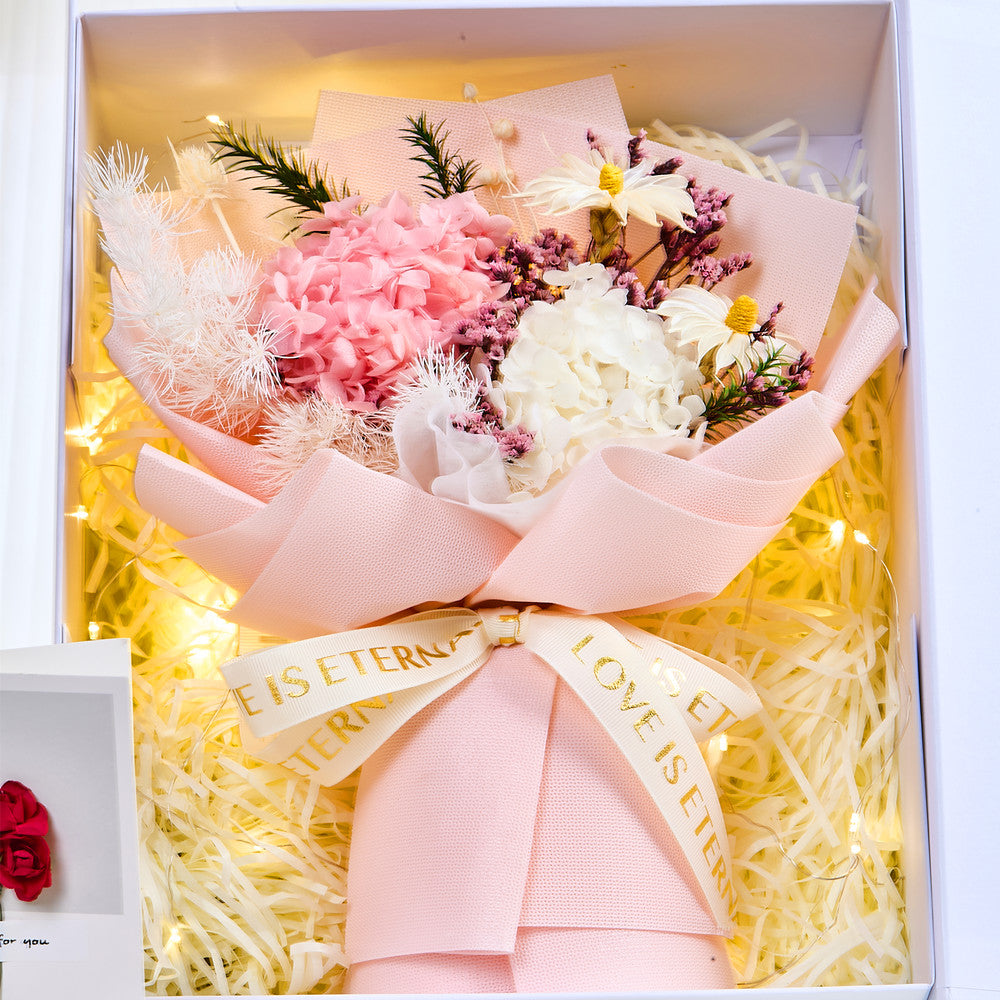 Top-down view of Eternal Glow Preserved Hydrangea Gift Box interior showing a pink and white preserved hydrangea bouquet with purple statice, dried daisies, and pampas grass, wrapped in blush pink paper with a Love is Eternal ribbon, nestled in cream shredded paper with warm fairy lights