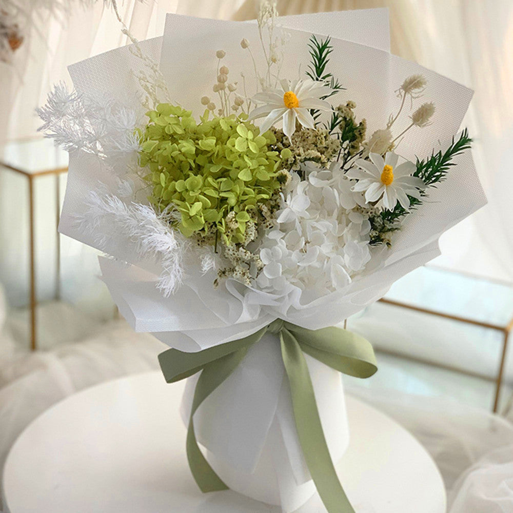 A dried flower bouquet wrapped in white tissue paper and tied with a sage green ribbon, displayed on a round white table in a softly lit indoor setting.