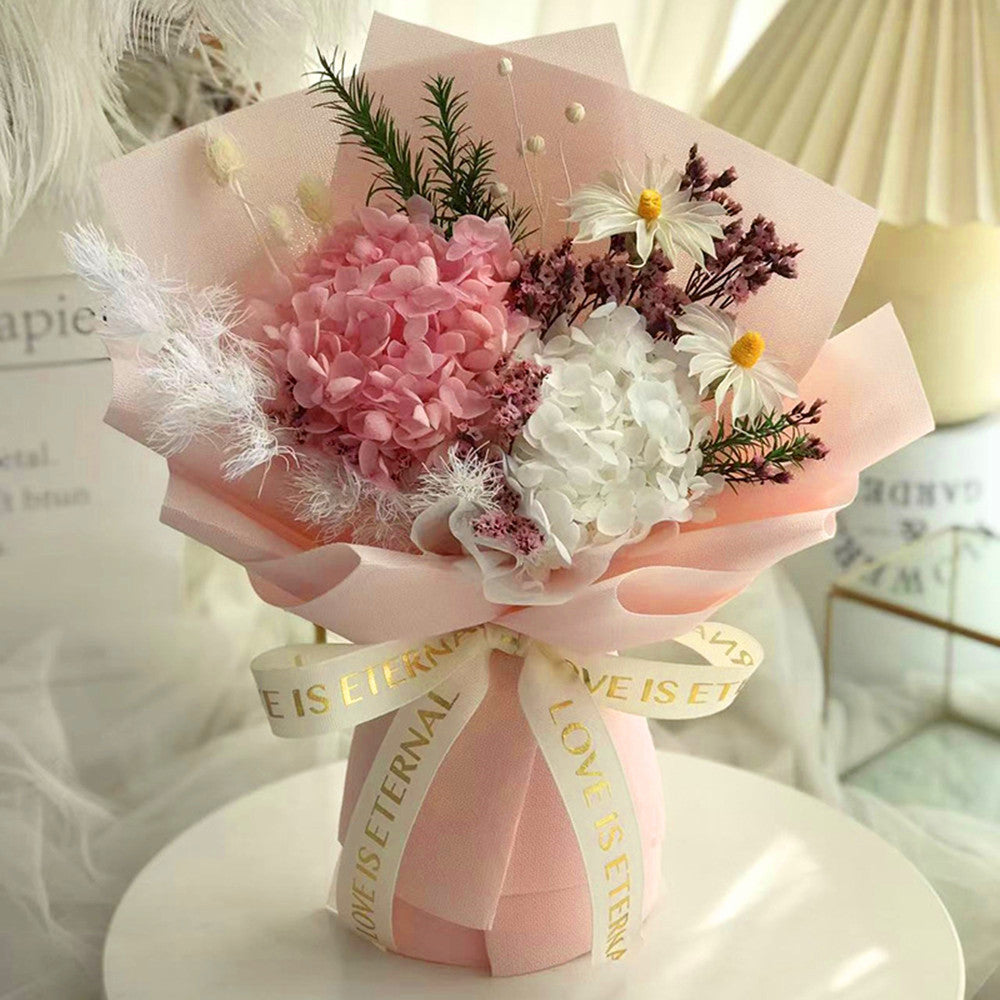 A decorative floral bouquet wrapped in pale pink paper with a matching ribbon printed with the words 'LOVE IS ETERNAL,' arranged with pink and white hydrangeas, daisies, and small dark purple filler flowers on a white round surface.