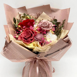 Front view of Bloom and Forever Eternal Glow Preserved Flower Gift Box Large bouquet, featuring red, deep pink, and mauve preserved roses with a dried sola flower, pink baby's breath, pink statice, cotton flower, and eucalyptus, wrapped in blush pink kraft paper with a mauve mesh ribbon, with warm fairy lights glowing throughout, on a white background