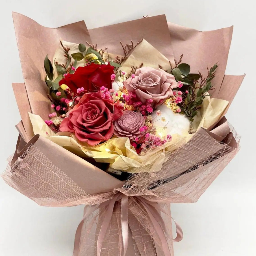 Front view of Bloom and Forever Eternal Glow Preserved Flower Gift Box Large bouquet, featuring red, deep pink, and mauve preserved roses with a dried sola flower, pink baby's breath, pink statice, cotton flower, and eucalyptus, wrapped in blush pink kraft paper with a mauve mesh ribbon, with warm fairy lights glowing throughout, on a white background