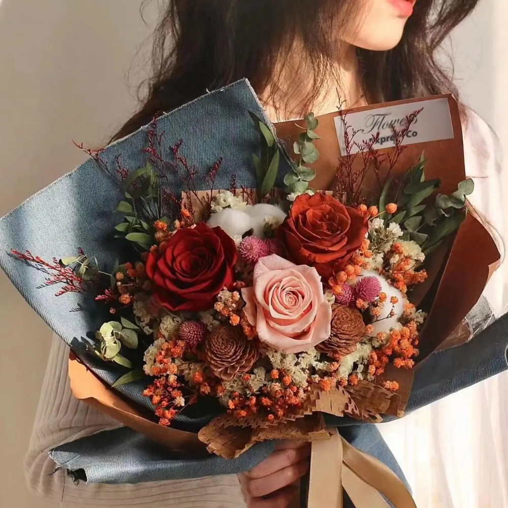 Woman holding the Bloom and Forever Eternal Glow Preserved Flower Gift Box Large bouquet, featuring red, burnt orange, and blush pink preserved roses with dried pine cones, orange hypericum berries, globe amaranth, cotton flowers, and eucalyptus, wrapped in denim blue and kraft paper with a tan ribbon, against a soft neutral background