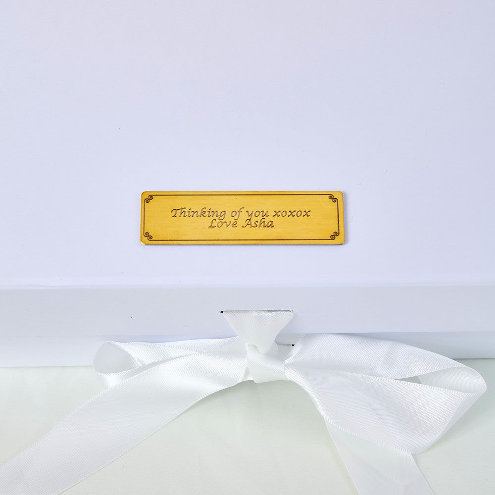 Close-up of a gold engraved personalised plaque on a white gift box lid reading Thinking of you xoxox Love Asha in elegant script, with a white satin ribbon bow below, highlighting the bespoke personalisation option for the Bloom and Forever Eternal Glow Preserved Flower Gift Box Large