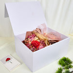 Side view of Bloom and Forever Eternal Glow Preserved Flower Gift Box Large open on a cream surface, showing a red preserved rose bouquet with pink baby's breath, pink statice, and dried fillers, wrapped in blush pink and kraft paper with a pink mesh ribbon, nestled inside a large white luxury gift box with warm fairy lights, with an Especially for You greeting card and green pine plant beside it