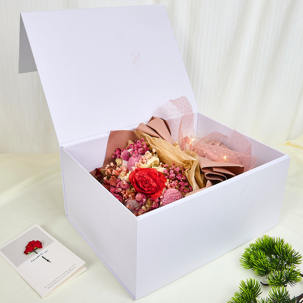 Side view of Bloom and Forever Eternal Glow Preserved Flower Gift Box Large open on a cream surface, showing a red preserved rose bouquet with pink baby's breath, pink statice, and dried fillers, wrapped in blush pink and kraft paper with a pink mesh ribbon, nestled inside a large white luxury gift box with warm fairy lights, with an Especially for You greeting card and green pine plant beside it