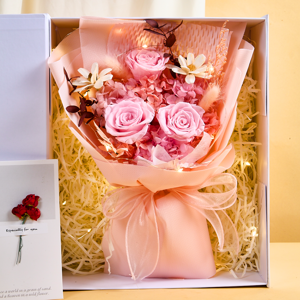 Top-down view inside the Bloom and Forever Eternal Glow Preserved Flower Gift Box Medium Blush Pink, showing three soft pink preserved roses with pink hydrangea, white dried daisies, bunny tail grass, and dark foliage, wrapped in blush pink paper with a peach organza ribbon, surrounded by cream shredded paper with warm glowing fairy lights, with an Especially for You greeting card visible