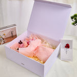 Side view of Bloom and Forever Eternal Glow Preserved Flower Gift Box Medium Blush Pink open on a white surface, showing a blush pink preserved rose bouquet wrapped in blush pink paper with a peach ribbon inside a white luxury gift box, with an Especially for You greeting card beside it and a green pine plant in the background