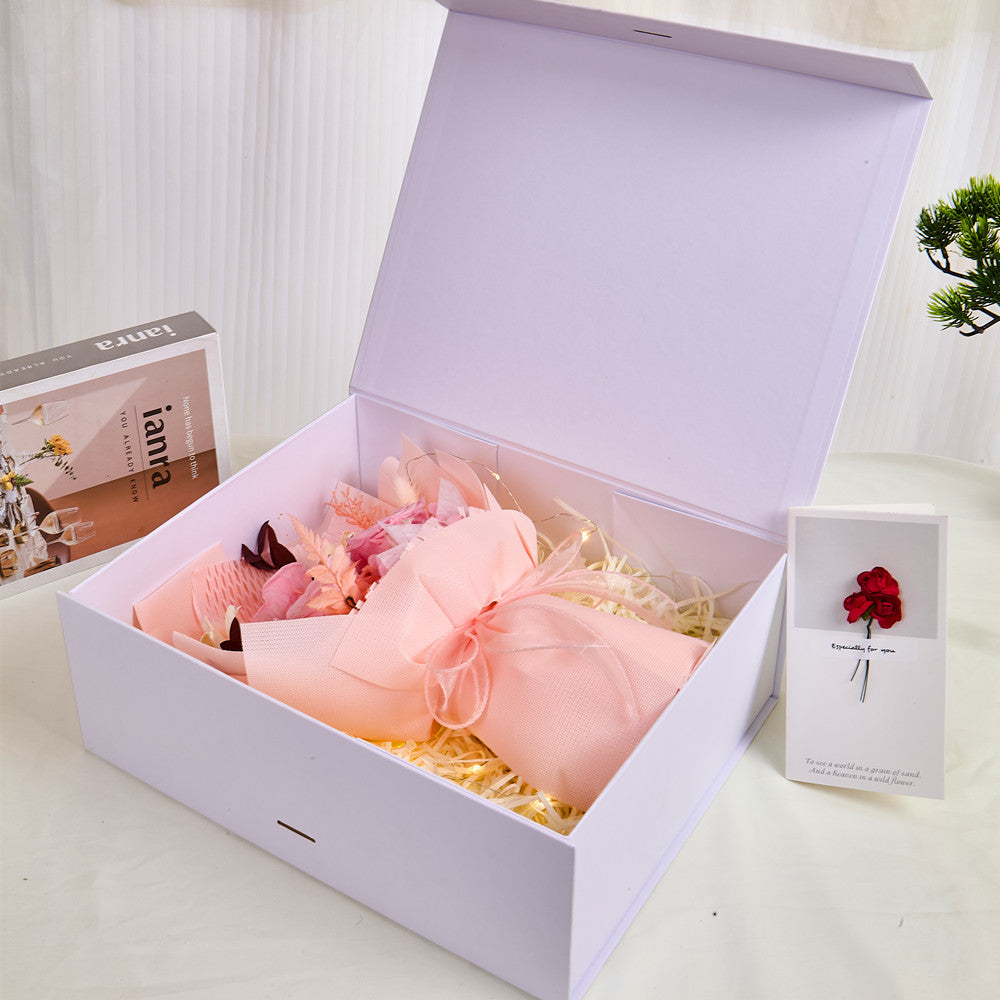 Side view of Bloom and Forever Eternal Glow Preserved Flower Gift Box Medium Blush Pink open on a white surface, showing a blush pink preserved rose bouquet wrapped in blush pink paper with a peach ribbon inside a white luxury gift box, with an Especially for You greeting card beside it and a green pine plant in the background