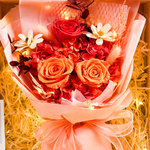 Close-up of Eternal Glow Preserved Flower Gift Box Medium Orange and Red interior, showing a vibrant bouquet of red and orange preserved roses with red hydrangea, white dried daisies, pink bunny tail grass, and dark foliage, wrapped in blush pink paper with a peach satin ribbon, nestled in cream shredded paper with warm glowing fairy lights