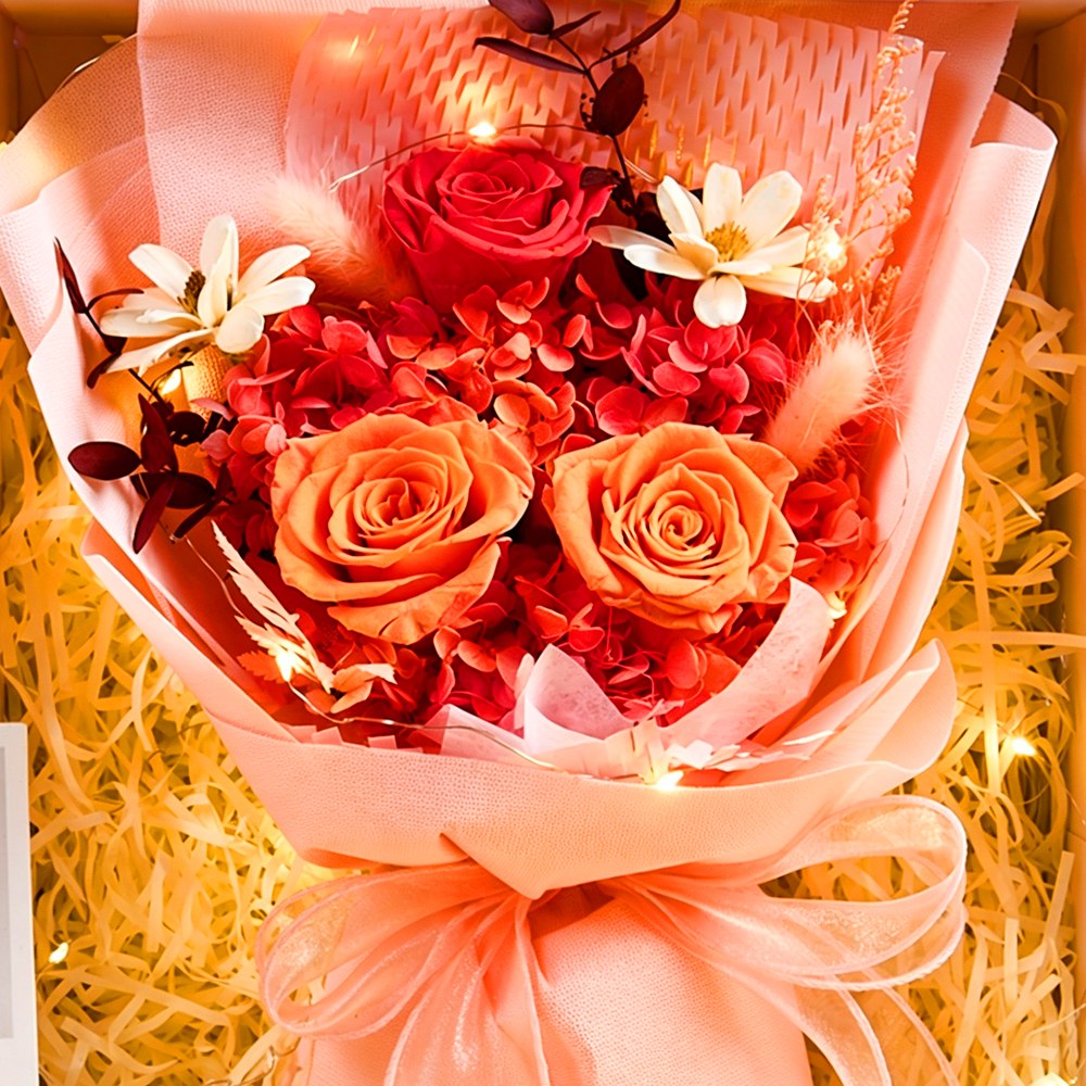 Close-up of Eternal Glow Preserved Flower Gift Box Medium Orange and Red interior, showing a vibrant bouquet of red and orange preserved roses with red hydrangea, white dried daisies, pink bunny tail grass, and dark foliage, wrapped in blush pink paper with a peach satin ribbon, nestled in cream shredded paper with warm glowing fairy lights