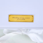 Close-up of a gold engraved personalised plaque on a white gift box lid reading Thinking of you xoxox Love Asha in elegant script, with a white satin ribbon bow below, highlighting the bespoke personalisation option for the Bloom and Forever Eternal Glow Preserved Flower Gift Box Medium Orange and Red