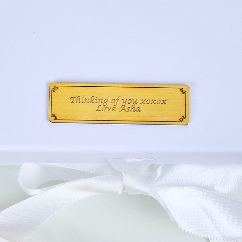 Close-up of a gold engraved personalised plaque on a white gift box lid reading Thinking of you xoxox Love Asha in elegant script, with a white satin ribbon bow below, highlighting the bespoke personalisation option for the Bloom and Forever Eternal Glow Preserved Flower Gift Box Medium Orange and Red