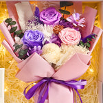 Close-up of Eternal Glow Preserved Flower Gift Box Medium Vintage Purple interior, showing a lush bouquet of purple, lavender, and pink preserved roses with white hydrangea, dried daisies, orange hypericum berries, purple statice, baby's breath, and eucalyptus, wrapped in blush pink paper with a purple satin ribbon, nestled in cream shredded paper with warm fairy lights
