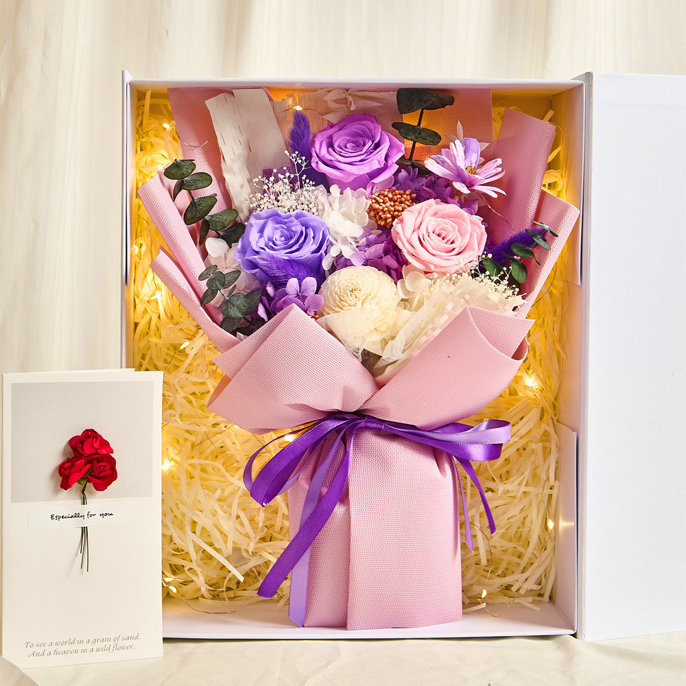 Top-down view inside the Bloom and Forever Eternal Glow Preserved Flower Gift Box Medium Vintage Purple, showing a purple, lavender, and pink preserved rose bouquet with white hydrangea, dried daisies, orange berries, and eucalyptus, wrapped in blush pink paper with a purple satin ribbon, surrounded by cream shredded paper and warm glowing fairy lights, with an Especially for You greeting card visible