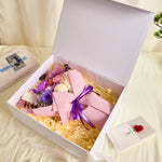 Angled overhead view of Bloom and Forever Eternal Glow Preserved Flower Gift Box Medium Vintage Purple open on a cream bed, showing a purple and pink preserved rose bouquet with blush pink wrapping and purple ribbon inside a white luxury gift box, with an Especially for You greeting card beside it and a magazine and pine plant in the background