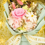 Close-up of Eternal Glow Preserved Flower Gift Box Small interior showing a pink preserved rose with white hydrangea, dried daisies, pink bunny tail grass, and white statice, wrapped in sage green floral paper with a Love is Eternal gold ribbon, nestled in cream shredded paper with warm fairy lights