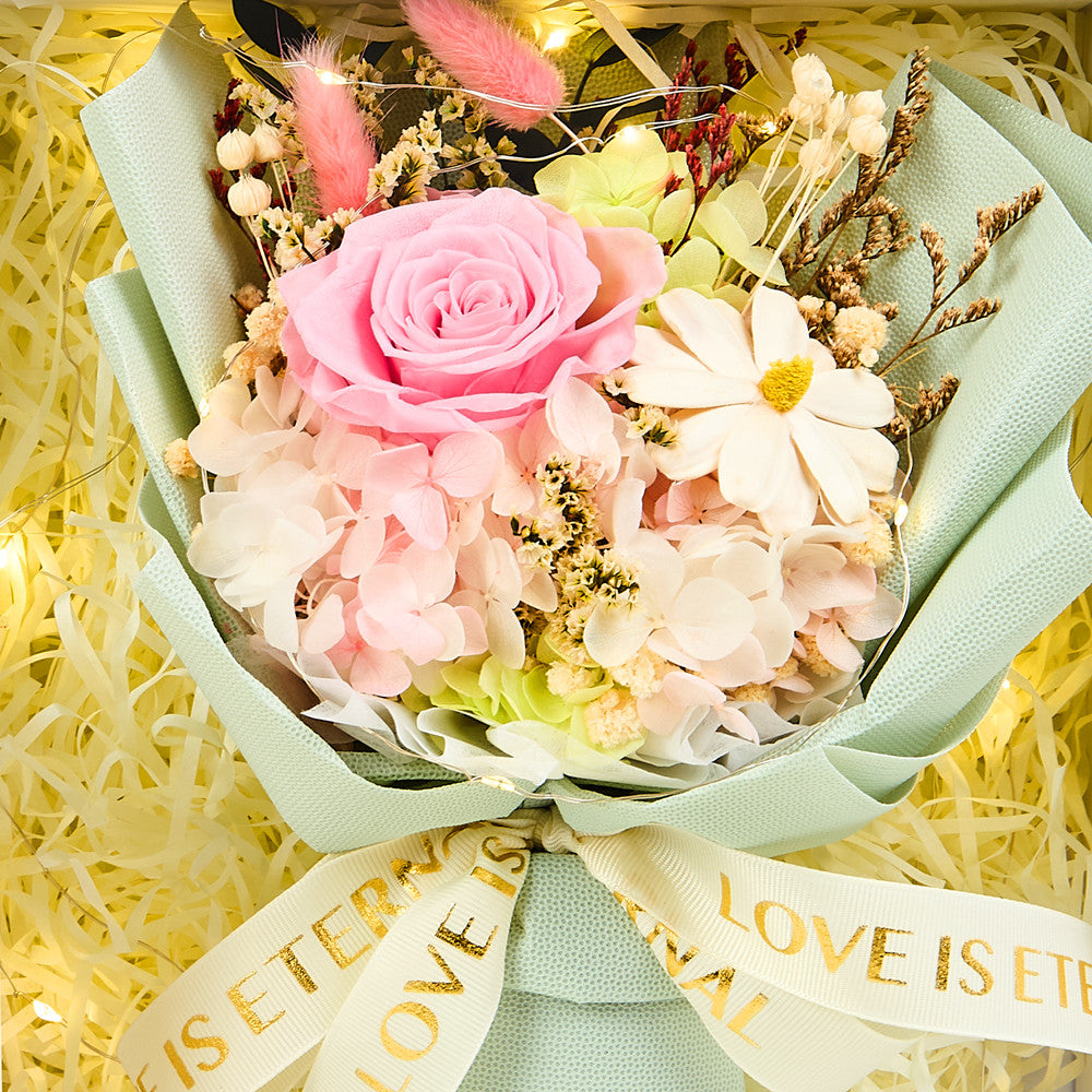 Close-up of Eternal Glow Preserved Flower Gift Box Small interior showing a pink preserved rose with white hydrangea, dried daisies, pink bunny tail grass, and white statice, wrapped in sage green floral paper with a Love is Eternal gold ribbon, nestled in cream shredded paper with warm fairy lights