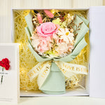 Top-down view inside the Eternal Glow Preserved Flower Gift Box Small by Bloom and Forever, showing a pink preserved rose and white hydrangea bouquet with dried daisies and bunny tail grass, wrapped in sage green paper with Love is Eternal ribbon, surrounded by cream shredded paper and warm fairy lights