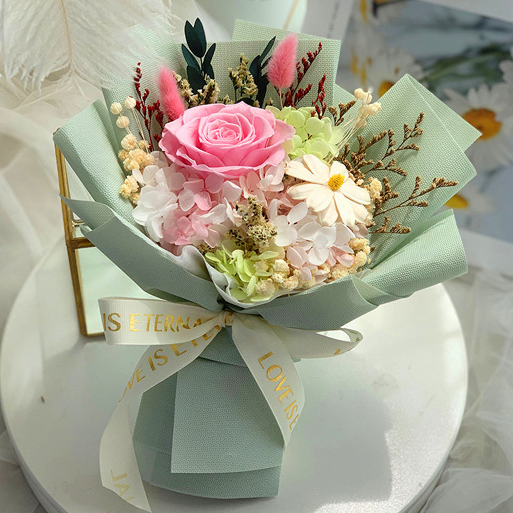A small floral bouquet wrapped in pale mint green paper with a white ribbon printed with gold text, arranged on a white round surface. The bouquet includes a pink rose, pink and white hydrangeas, small cream filler flowers, and green foliage.