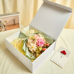 Side view of Bloom and Forever Eternal Glow Preserved Flower Gift Box Small open on a cream surface, showing a pink preserved rose bouquet wrapped in sage green paper with Love is Eternal ribbon inside a white luxury gift box, with an Especially for You greeting card beside it