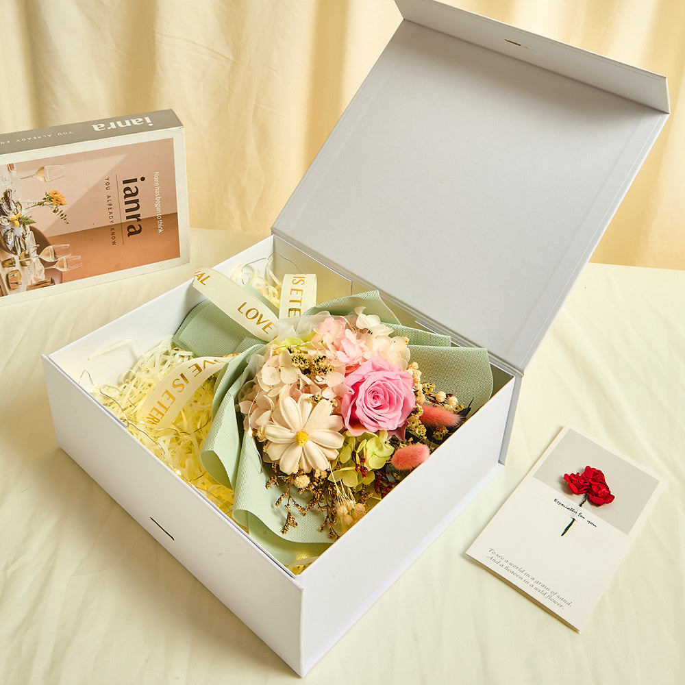 Side view of Bloom and Forever Eternal Glow Preserved Flower Gift Box Small open on a cream surface, showing a pink preserved rose bouquet wrapped in sage green paper with Love is Eternal ribbon inside a white luxury gift box, with an Especially for You greeting card beside it