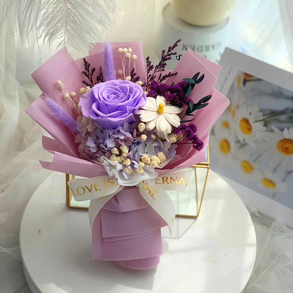 A small decorative bouquet wrapped in pink paper, featuring a lavender rose, white daisy, purple filler flowers, and dried botanicals, tied with a white ribbon that reads 'LOVE IS.