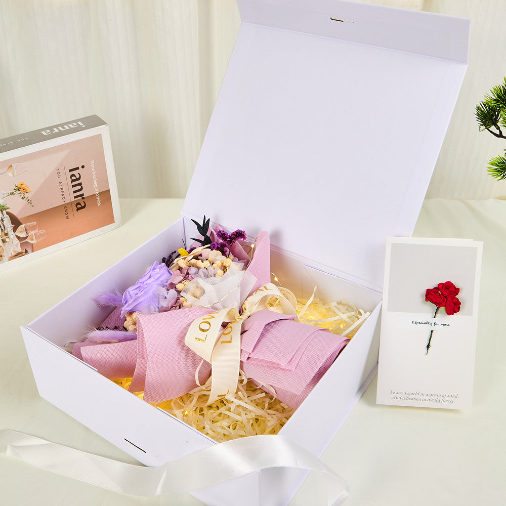 Side view of Bloom and Forever Eternal Glow Preserved Flower Gift Box Small open on a cream surface, showing a purple preserved rose bouquet wrapped in blush pink paper with Love is Eternal ribbon inside a white luxury gift box, with an Especially for You greeting card beside it