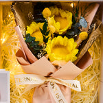 Top-down close-up of Eternal Glow Preserved Flower Gift Box Sunflower interior, showing a vibrant yellow preserved sunflower bouquet with craspedia billy balls, eucalyptus, and dried fillers, wrapped in kraft paper with a Love is Eternal ribbon, nestled in cream shredded paper with warm glowing fairy lights