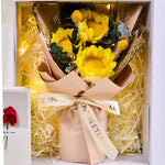 Top-down view inside the Eternal Glow Preserved Flower Gift Box Sunflower by Bloom and Forever, showing a yellow preserved sunflower and craspedia bouquet with eucalyptus and dried fillers, wrapped in kraft paper with Love is Eternal ribbon, surrounded by cream shredded paper and warm fairy lights, with an Especially for You greeting card visible