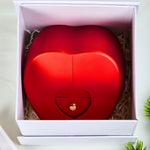 Top-down view of the red Bloom and Forever Eternal Rose Apple Jewelry Gift Box nestled inside a white outer gift box with cream shredded paper, showing the closed apple-shaped box with a gold heart drawer pull, with a green plant visible in the corner