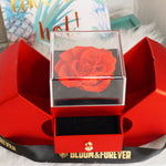 Lifestyle shot of Bloom and Forever Eternal Rose Apple Jewelry Gift Box in red with apple-shaped doors open, displaying a red preserved rose under a clear acrylic cover on a black velvet base with the jewelry drawer pulled out, placed on a red surface with Bloom and Forever branded ribbon and a tropical print background