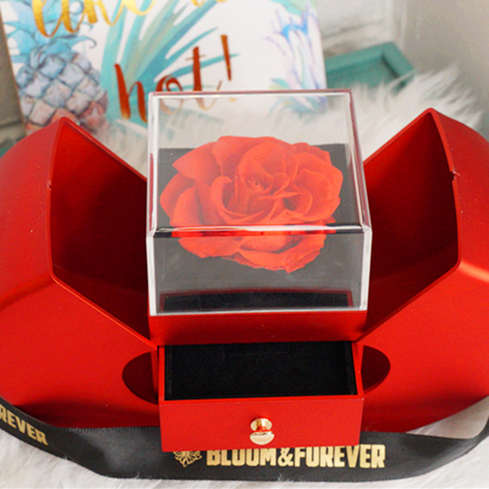 Lifestyle shot of Bloom and Forever Eternal Rose Apple Jewelry Gift Box in red with apple-shaped doors open, displaying a red preserved rose under a clear acrylic cover on a black velvet base with the jewelry drawer pulled out, placed on a red surface with Bloom and Forever branded ribbon and a tropical print background