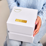 Person in a blue striped shirt holding the closed white outer gift box for the Bloom and Forever Eternal Rose Apple Jewelry Gift Box, showing the Bloom and Forever logo and bloomforever.com.au branding in gold with a personalised gold engraved plaque reading Thinking of you xoxox Love Asha
