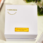Top-down view of the closed white outer gift box for the Bloom and Forever Eternal Rose Apple Jewelry Gift Box, showing the Bloom and Forever logo and bloomforever.com.au branding in gold with a personalised gold engraved plaque reading Thinking of you xoxox Love Asha, displayed on a white surface with a green plant in the corner