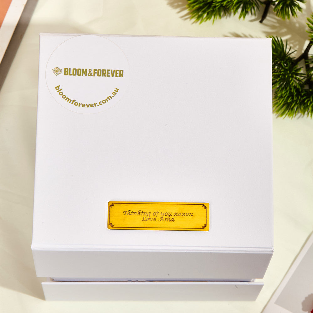 Top-down view of the closed white outer gift box for the Bloom and Forever Eternal Rose Apple Jewelry Gift Box, showing the Bloom and Forever logo and bloomforever.com.au branding in gold with a personalised gold engraved plaque reading Thinking of you xoxox Love Asha, displayed on a white surface with a green plant in the corner