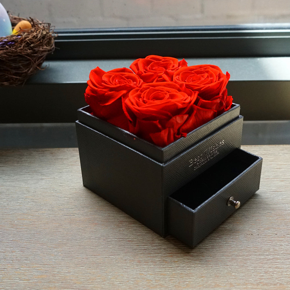 Bloom and Forever Eternal Rose Jewelry Drawer Gift Box in black leatherette with the jewelry drawer pulled open, displaying four vibrant red preserved roses on top, placed on a wooden surface with a decorative bird nest in the background