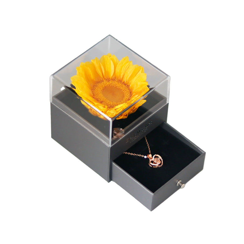 Bloom and Forever Eternal Sunflower Jewelry Drawer Box in grey with clear acrylic top, showing a vibrant yellow preserved sunflower under the lid and the jewelry drawer pulled open revealing a rose gold heart necklace inside the black velvet-lined drawer, on a white background