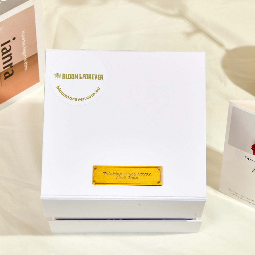 Top-down view of the closed white outer gift box for the Bloom and Forever Eternal Sunflower Jewelry Drawer Box, showing the Bloom and Forever logo and bloomforever.com.au branding in gold with a personalised gold engraved plaque reading Thinking of you xoxox Love Asha, displayed on a cream surface with an Especially for You greeting card and magazine beside it