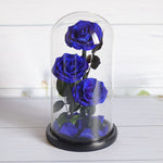 Three preserved royal blue roses with dark green leaves and stems, displayed inside a clear glass dome cloche with a black base on a white wooden surface.