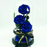 Three preserved royal blue roses with green leaves displayed under a clear glass dome on a black base, tied with a black ribbon printed with gold text.
