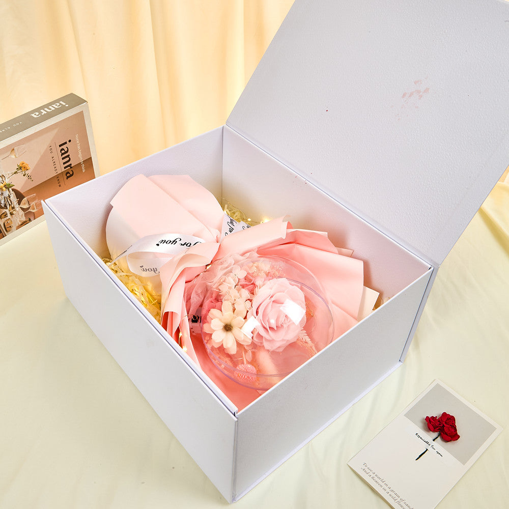 Side view of Bloom & Forever Fairy Light Preserved Flower Globe Gift Box open, showing a pink preserved rose globe bouquet wrapped in pink tissue with "Just for you" ribbon, sitting in a white luxury gift box with shredded paper filler and a greeting card beside it