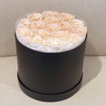 A round black cylindrical gift box filled with preserved pale peach roses, photographed against a neutral beige background.