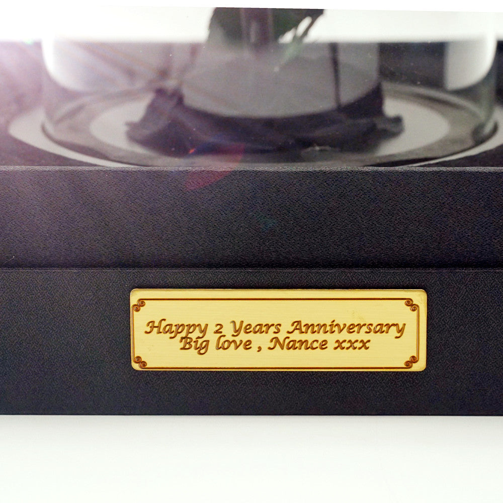 Close-up of a gold engraved personalised plaque on the black velvet base of a Bloom and Forever Everlasting Dome, displaying the custom message Happy 2 Years Anniversary Big love Nance xxx in elegant script, showcasing the bespoke personalisation option available for the Blush Hydrangea Everlasting Dome