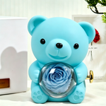 Bloom and Forever Rotating Eternal Rose Bear Gift Box in blue, front view showing a blue preserved rose encased in a clear heart-shaped acrylic globe held by the bear, with a gold wind-up key on the side, displayed beside a white outer gift box with personalised gold plaque on a cream surface