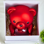 Top-down view of the red Bloom and Forever Rotating Eternal Rose Bear nestled inside a white outer gift box with cream shredded paper, showing the bear holding a red preserved rose in a clear heart-shaped acrylic globe with a gold wind-up key on the side