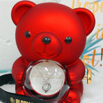 Close-up front view of the red Bloom and Forever Rotating Eternal Rose Bear with the clear heart-shaped acrylic globe rotated open, revealing a silver crystal drop necklace displayed on white velvet inside the jewelry compartment, with Bloom and Forever branded ribbon in the foreground